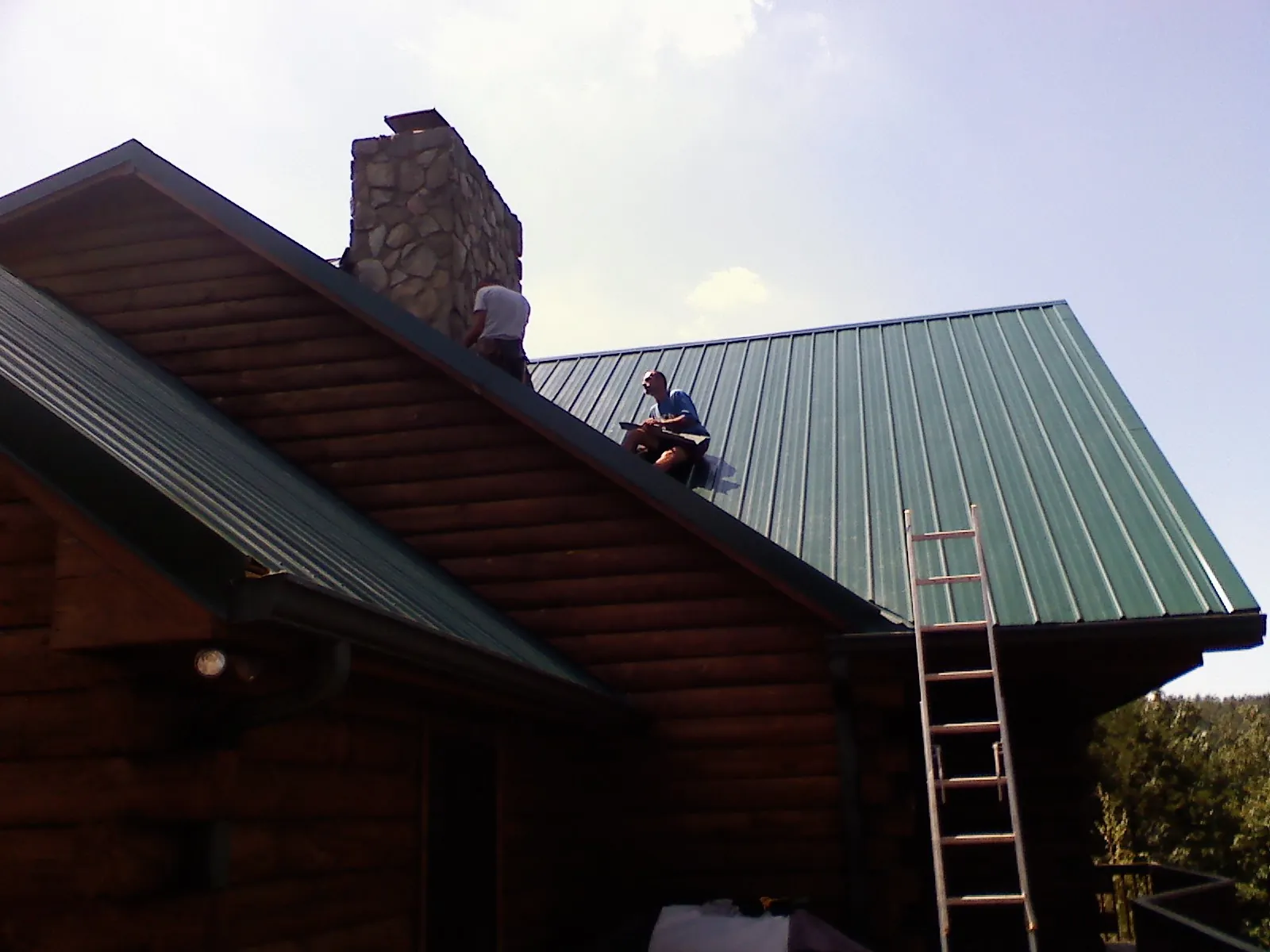 Roofers installing green metal panels beside a stone chimney on a log home in Collinsville